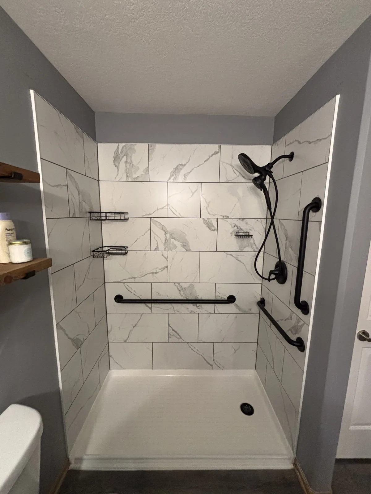Walk-in shower with large-format marble-look tile walls, a white base pan, black grab bars, and a handheld shower fixture