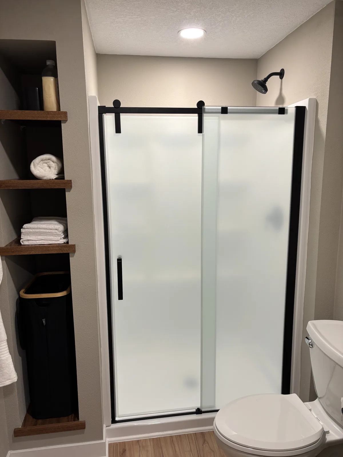 Shower with a frosted glass sliding door on black hardware beside a built-in linen nook with stained wood shelves