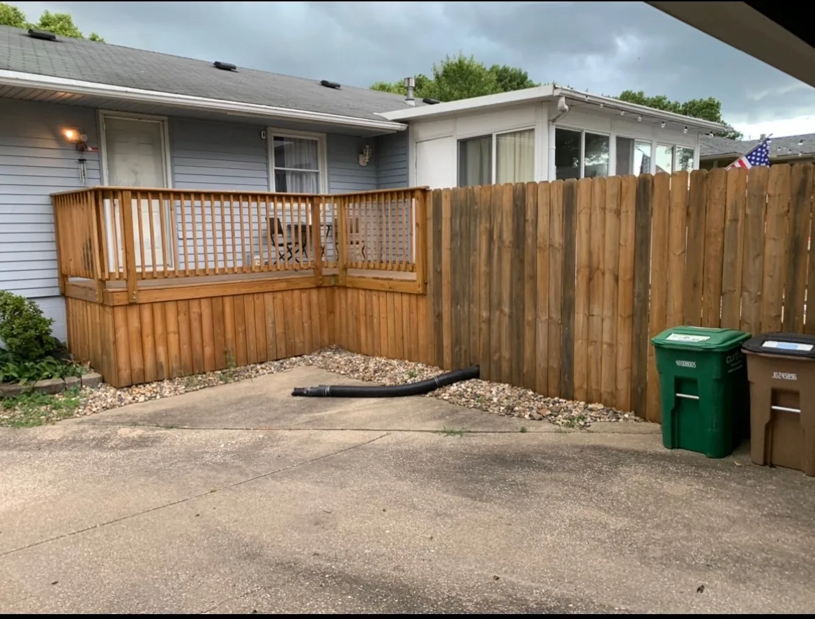 Concrete patio with stained deck railing, privacy fence, and river rock border along the house