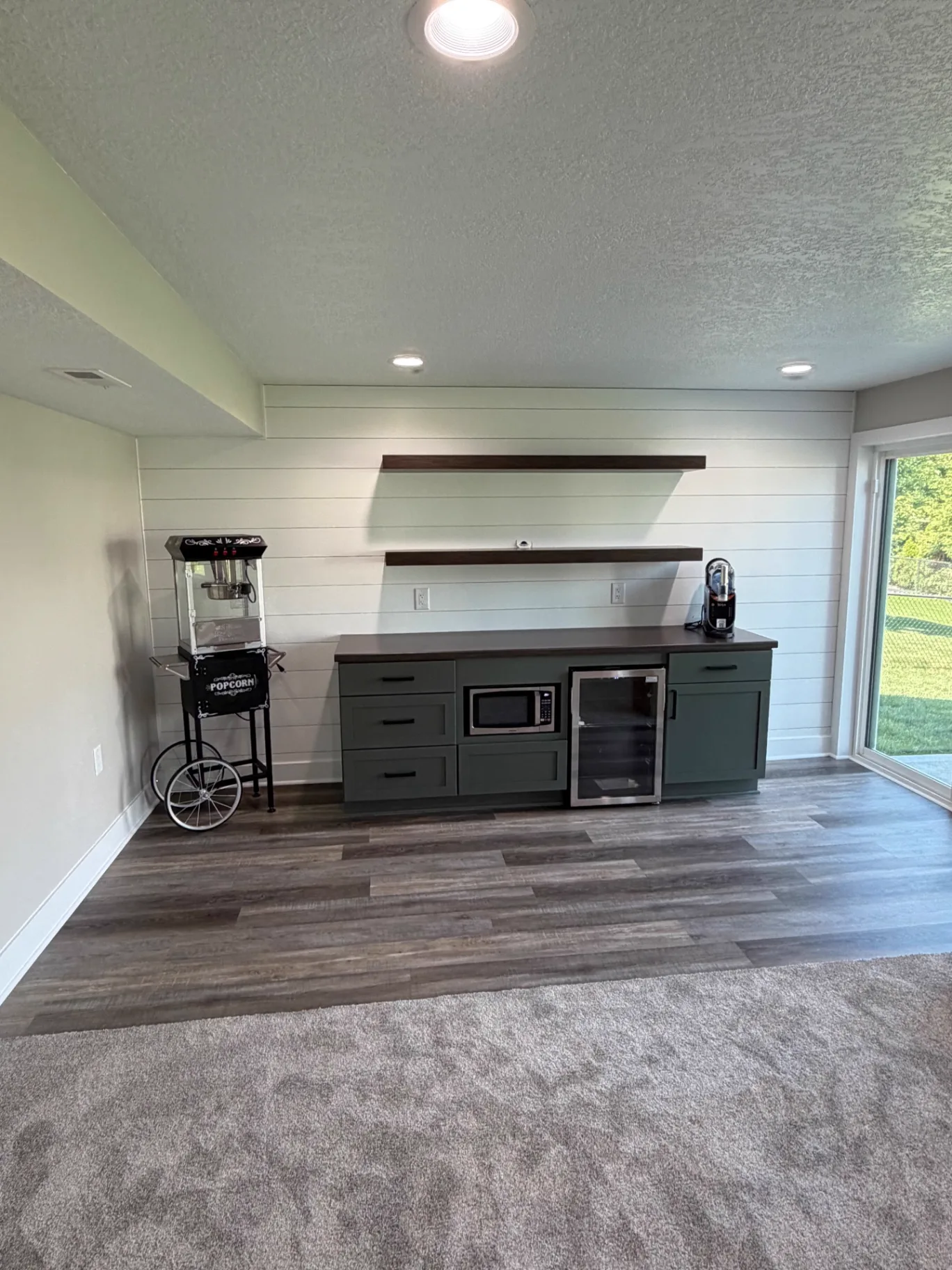 Basement snack and beverage bar with dark green cabinets, shiplap wall, floating wood shelves, and beverage fridge