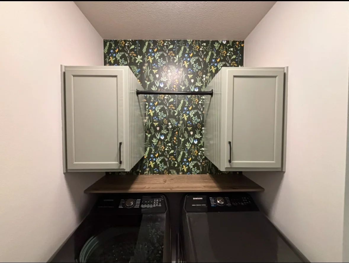 Laundry nook with sage green upper cabinets, botanical wallpaper, butcher block folding counter, and a black hanging rod