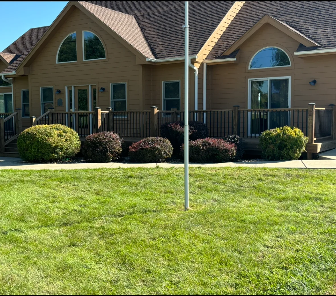 Front elevation of a tan home with a full-width wood deck and black metal railing behind landscaped shrubs