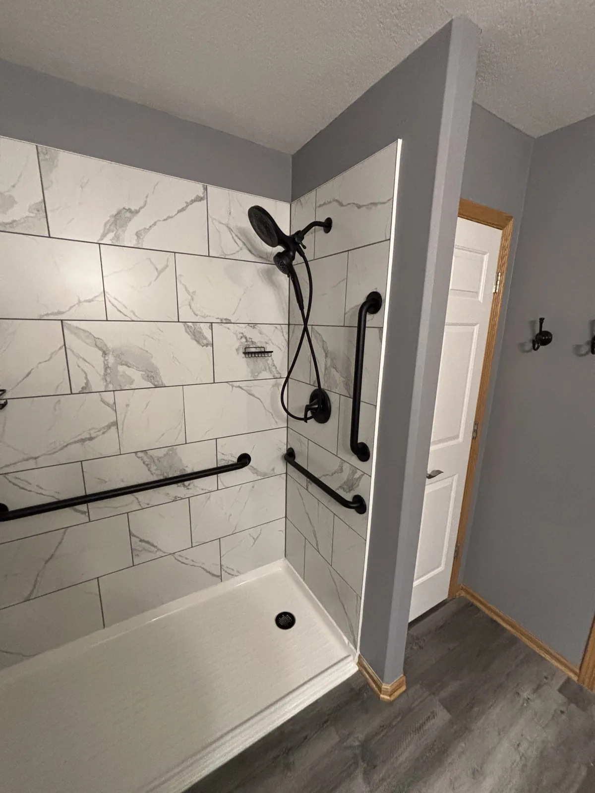 Angled view of a finished walk-in shower with marble-look tile, black grab bars, and wood-look vinyl plank flooring
