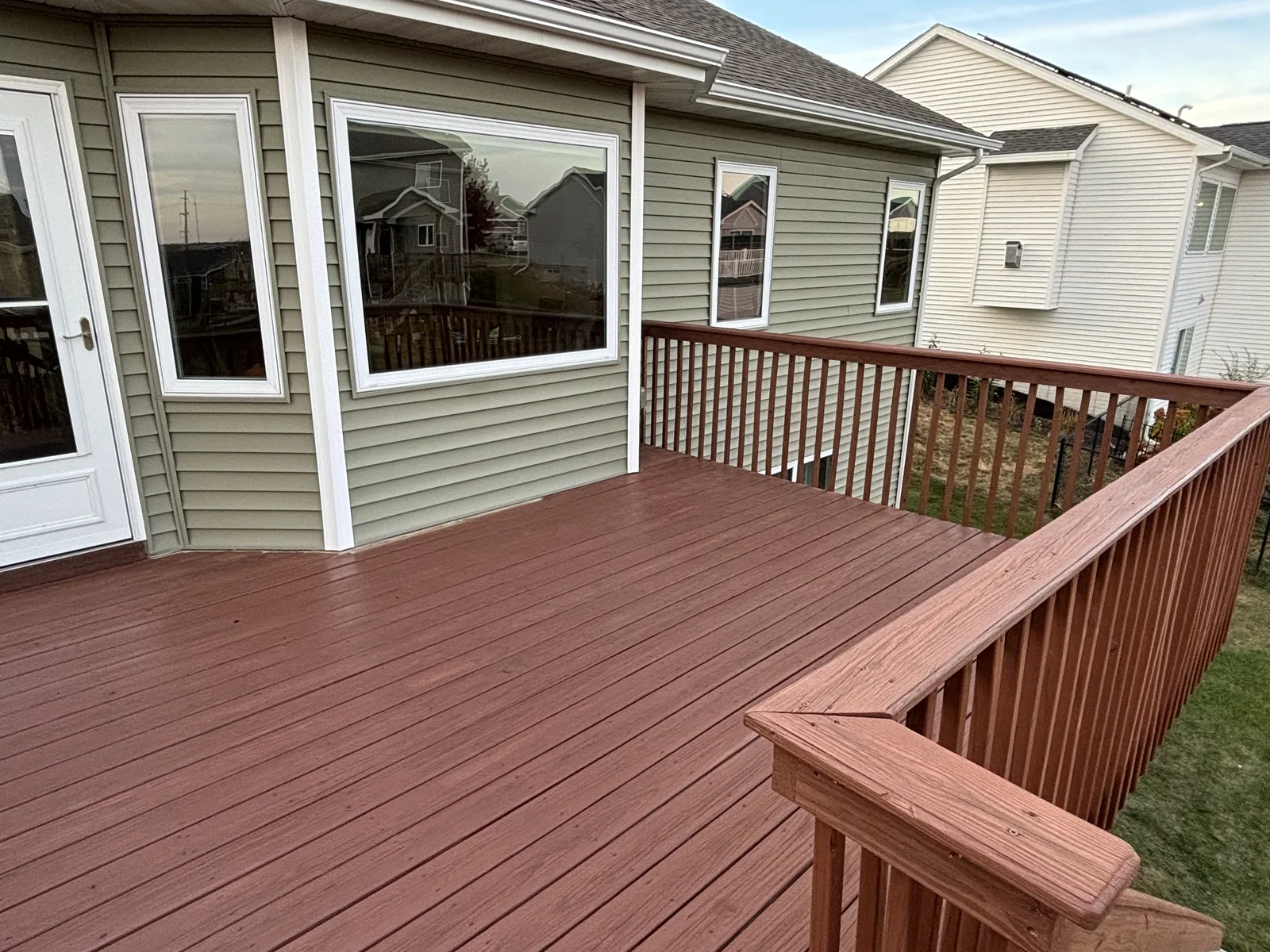 Freshly stained red-brown deck with matching railing along the back of a sage green sided home