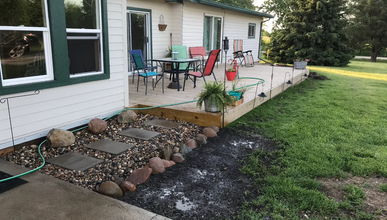 Ground-level deck with fresh dirt work, rock border, and stepping stone path along the side of a home