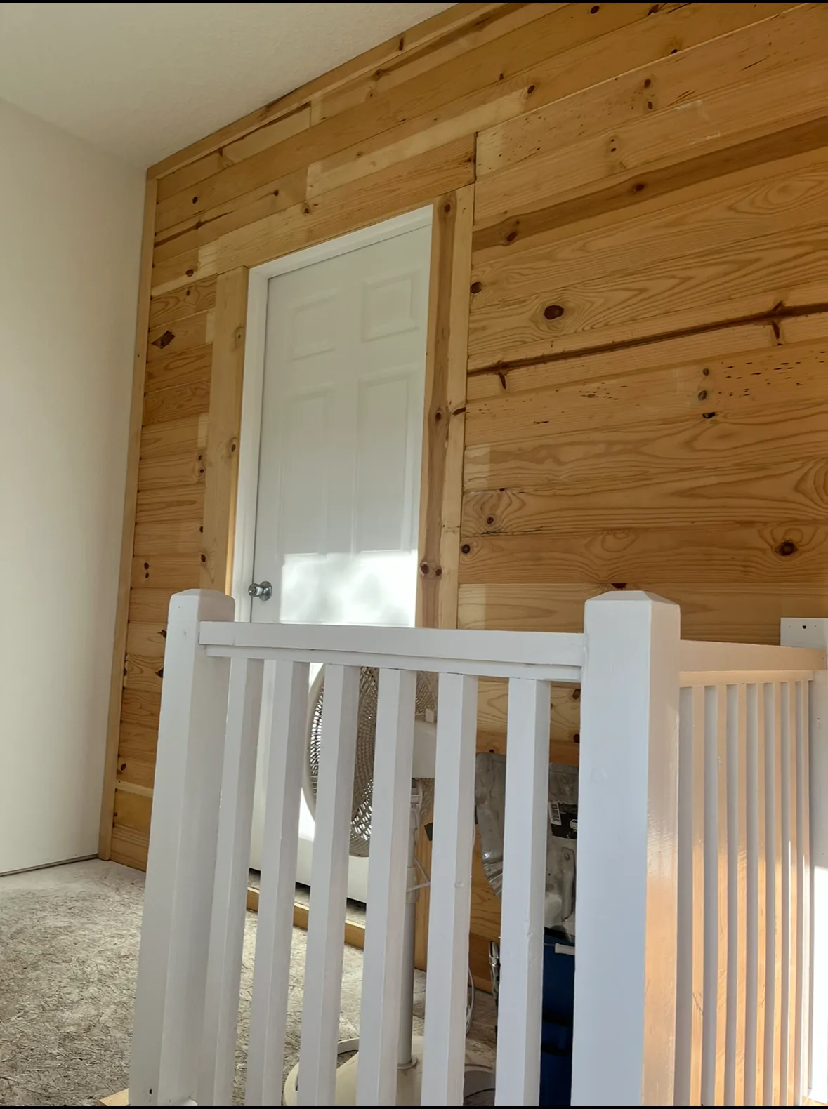 Interior wall clad in natural pine planking around a white six-panel door with a white stair railing in the foreground