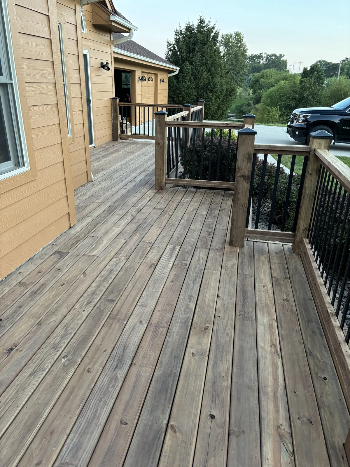 Wraparound wood deck with railing and black balusters running along the side of a tan house toward the garage