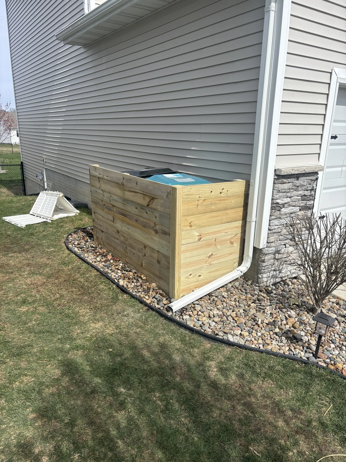 Pressure-treated wood screen enclosure built around a utility unit beside a home, set on a rock border