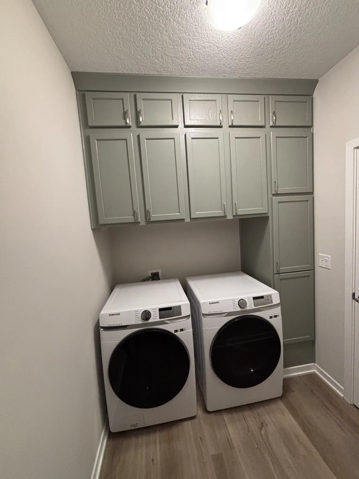 Laundry room with refinished sage green cabinetry above a washer and dryer and wood-look plank flooring